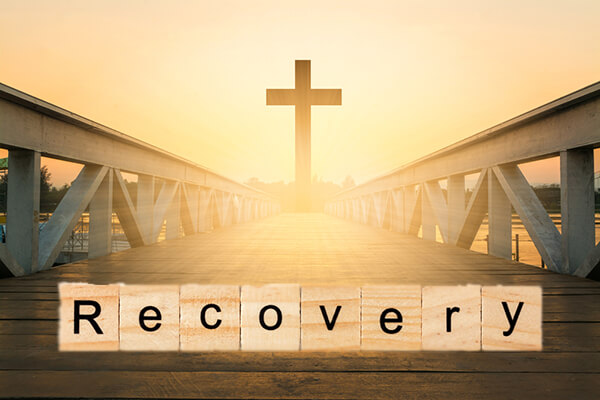 Bridge over water with cross in the middle with sun shining through and word "recovery" written over bridge